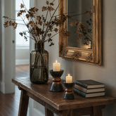 A styled interior scene showing a rustic wooden console table with a dark amber glass vase holding dried branches, two lit white pillar candles in dark candle holders, a stack of three books, and a gold-framed mirror on a beige wall.