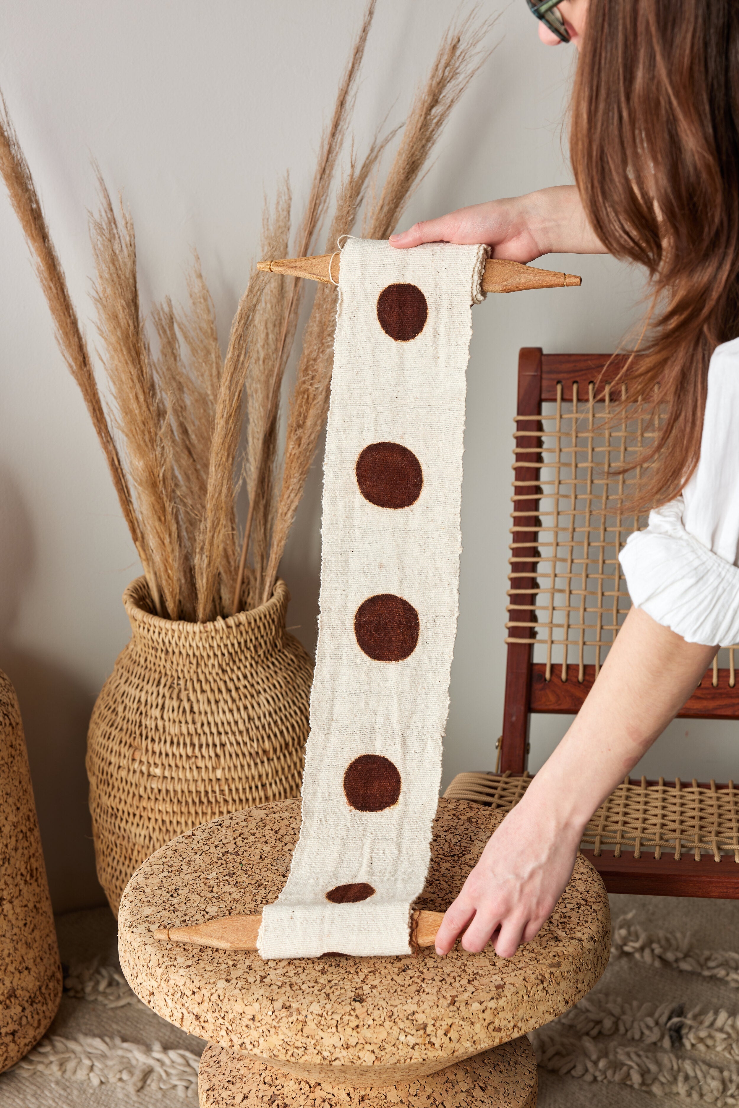 Person holding a decorative item with brown circles on a woven stool with pampas grass in the background.