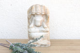 Stone sculpture of a seated figure on a wooden surface with a white textured wall background