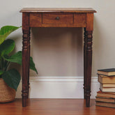 Wooden side table with a drawer next to a stack of books and a plant on a wooden floor.