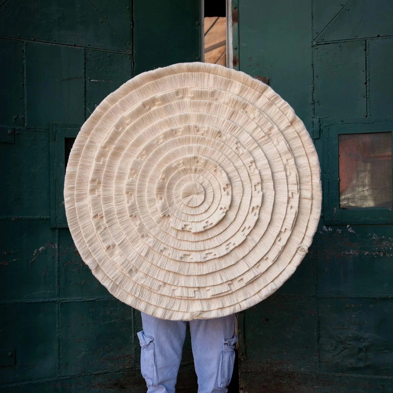 Person holding a large round woven mat against a dark textured wall