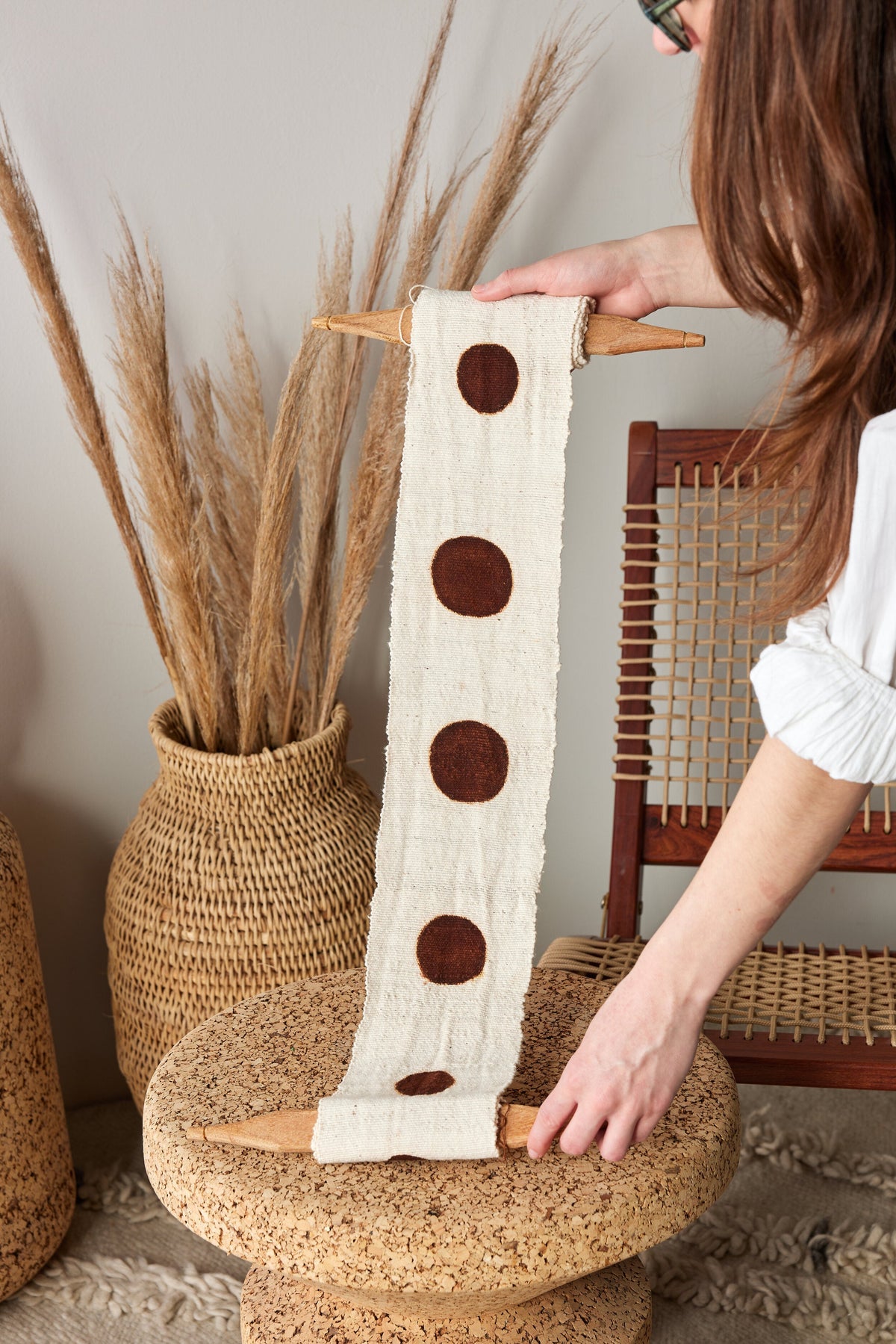 Person holding a decorative item with brown circles on a woven stool with pampas grass in the background.