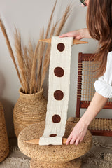 Person holding a decorative item with brown circles on a woven stool with pampas grass in the background.