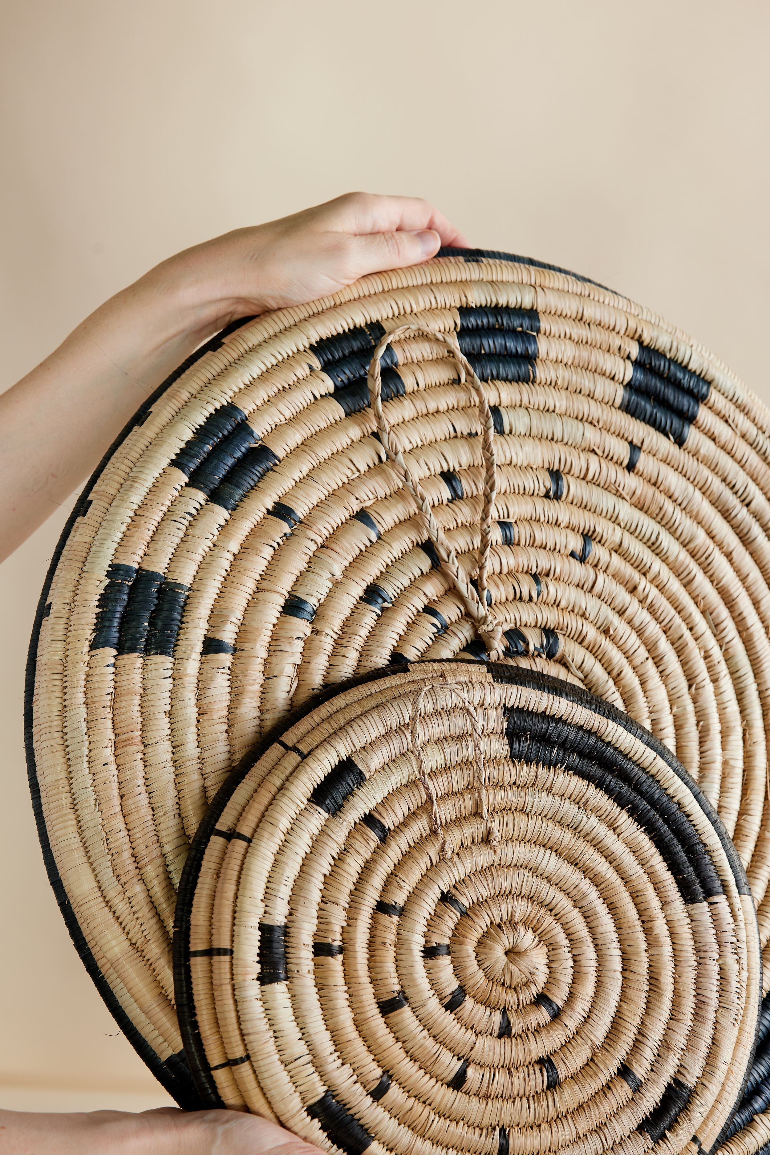 Hand holding two woven round trays with black patterns on a beige background