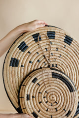 Hand holding two woven round trays with black patterns on a beige background