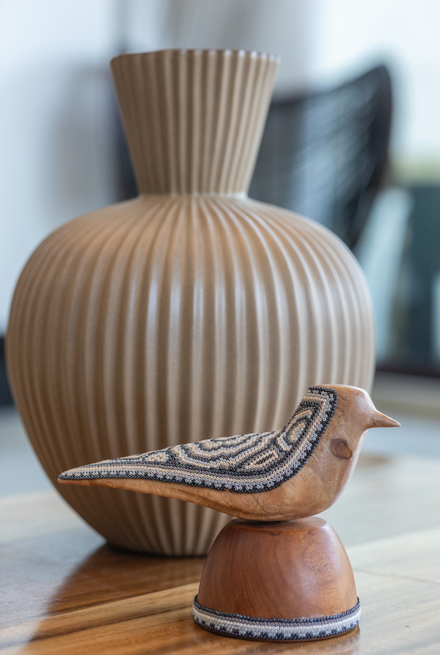 Wooden bird sculpture on a small pedestal in front of a ribbed beige vase.