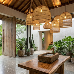 A modern rustic interior with multiple woven wicker pendant lights hanging over a wooden table, surrounded by potted plants and a stone water basin, in a tropical-style pavilion.