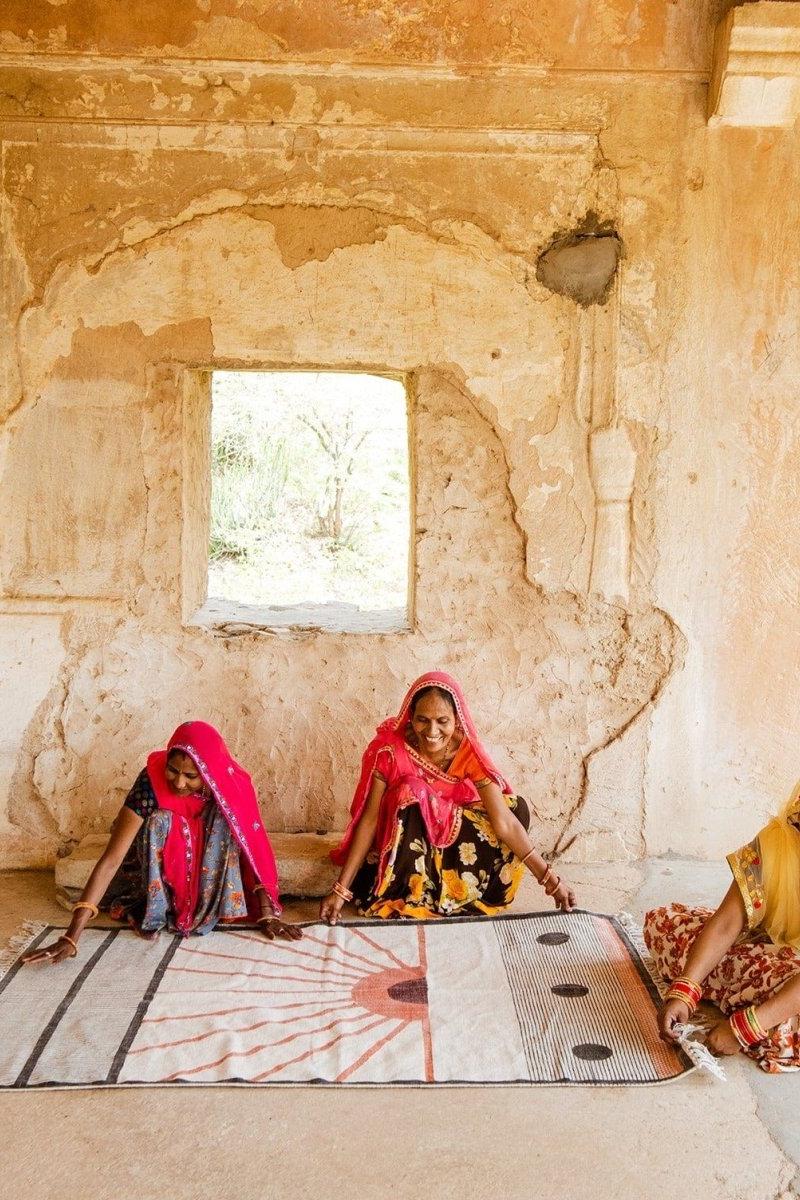 Three women in colorful traditional attire sitting on a patterned mat inside a rustic building.