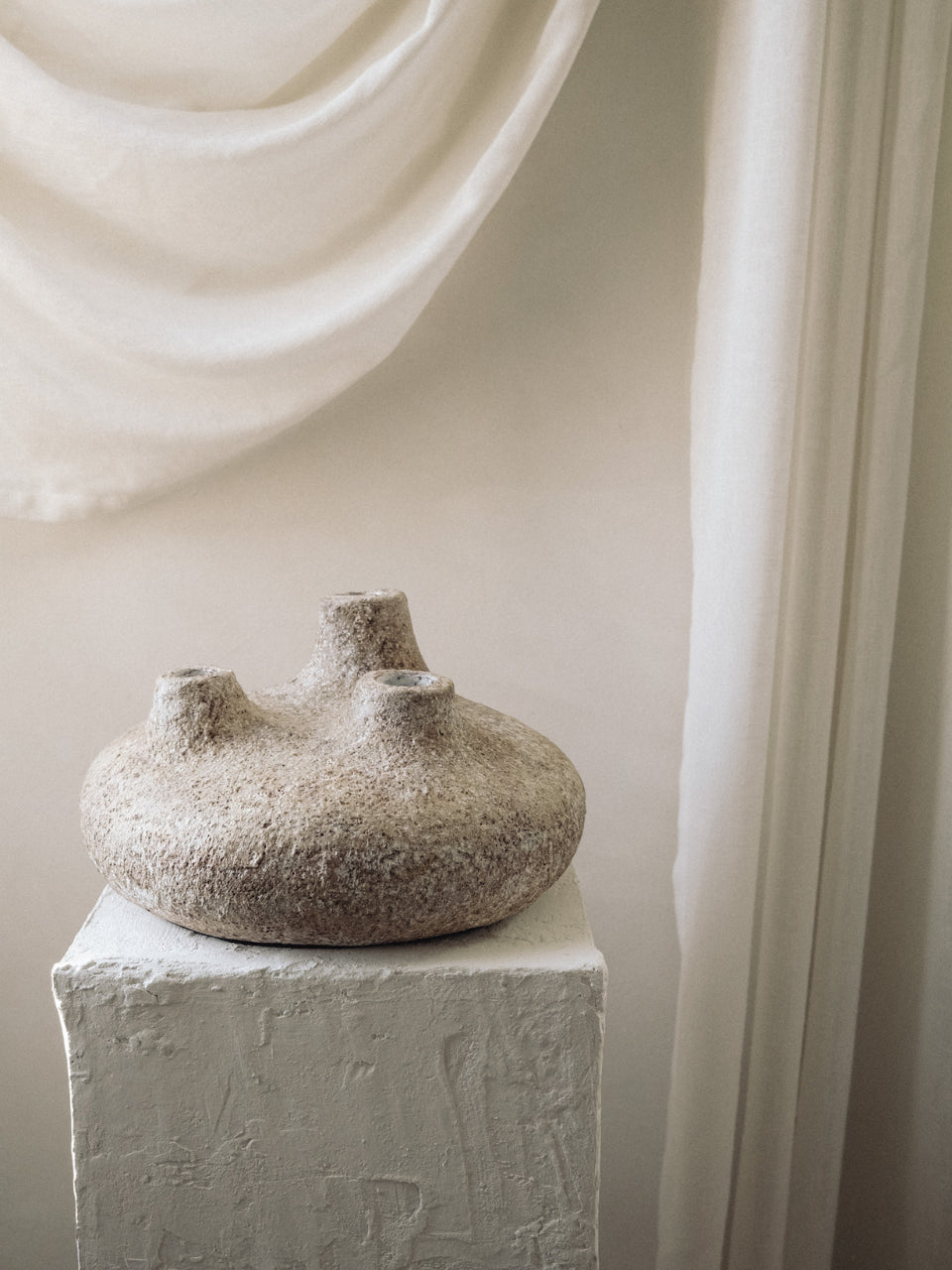 Textured vase on a white pedestal against a light curtain backdrop