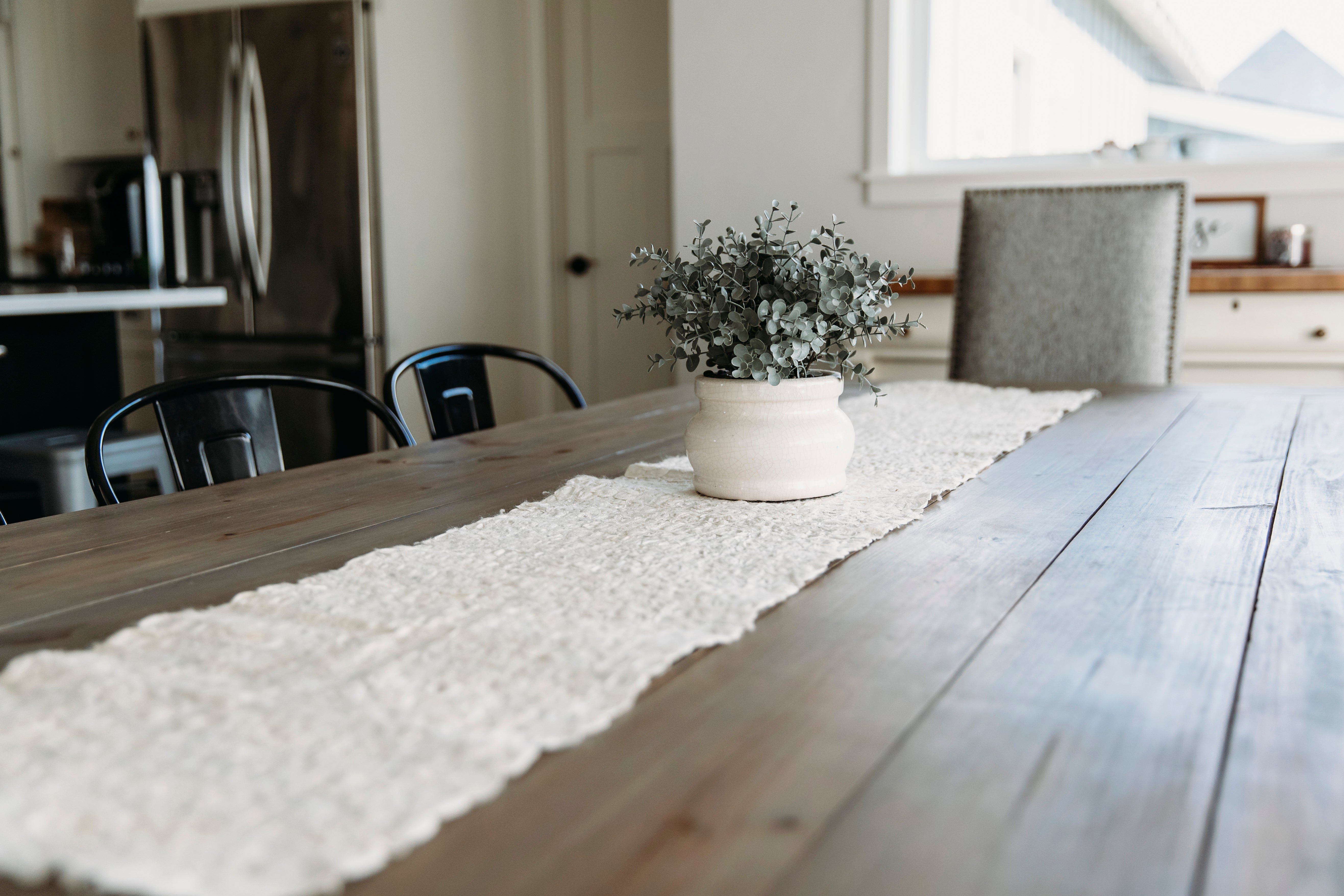 Long wooden dining table with a white textured runner and a small plant in the center, set against a kitchen background.